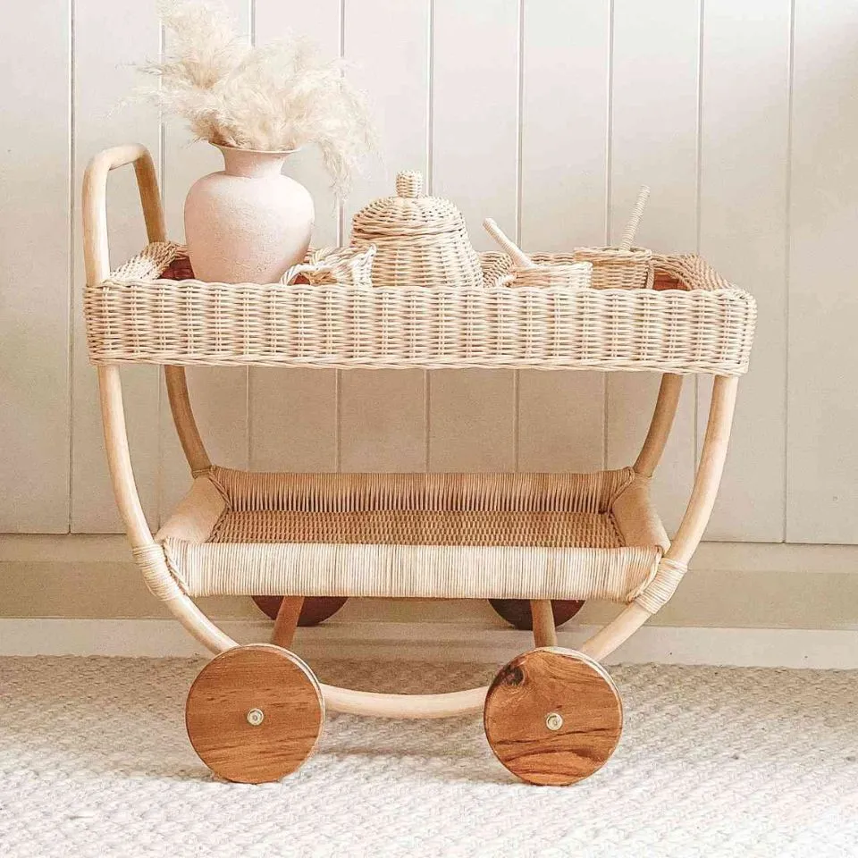 Handwoven rattan two-tier kids serving cart with wooden wheels, carrying teapot and vase – role-play essential.
